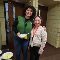 Two people standing in a hallway, smiling and holding bowls at a food service table.