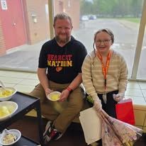 Two people seated indoors, smiling and holding dessert bowls near a window.