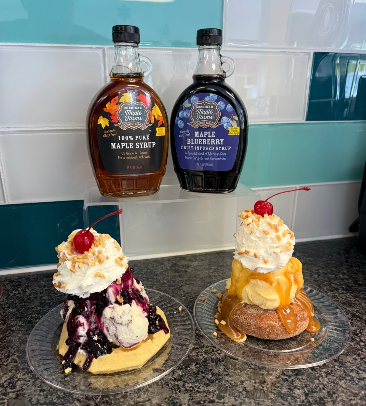 Two maple syrup bottles behind two ice cream sundaes with whipped cream and cherries on a counter.