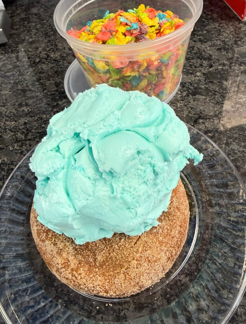 A blue ice cream scoop on a cinnamon-sugar coated dessert with a colorful candy cup behind it