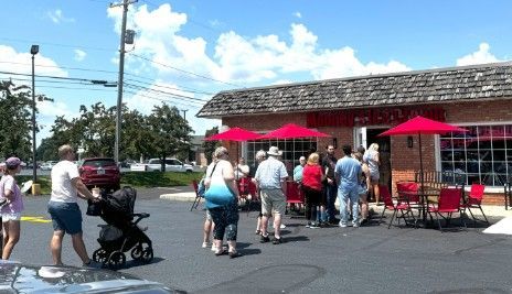 People standing in line outside a small building with red umbrellas on a sunny day