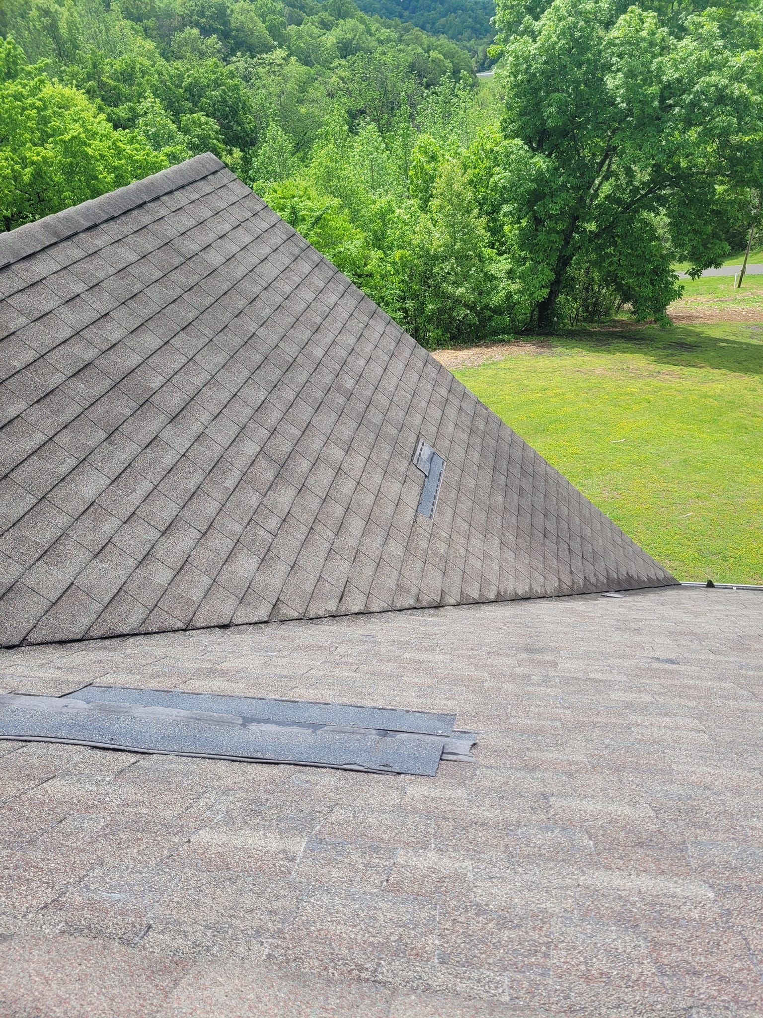 Close-up of a house roof, showing gray shingles with a patch of new shingles in the foreground. Green trees in the background.