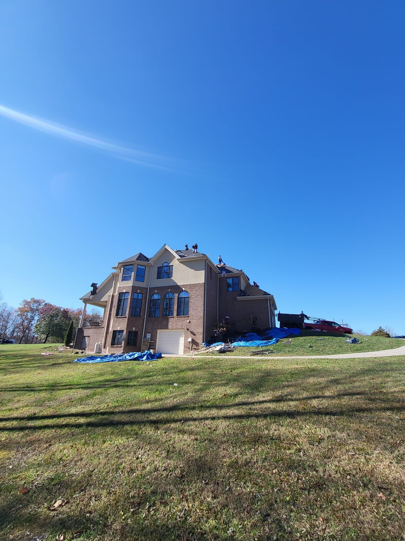 House under construction on a grassy hill, surrounded by blue tarps, against a clear blue sky.