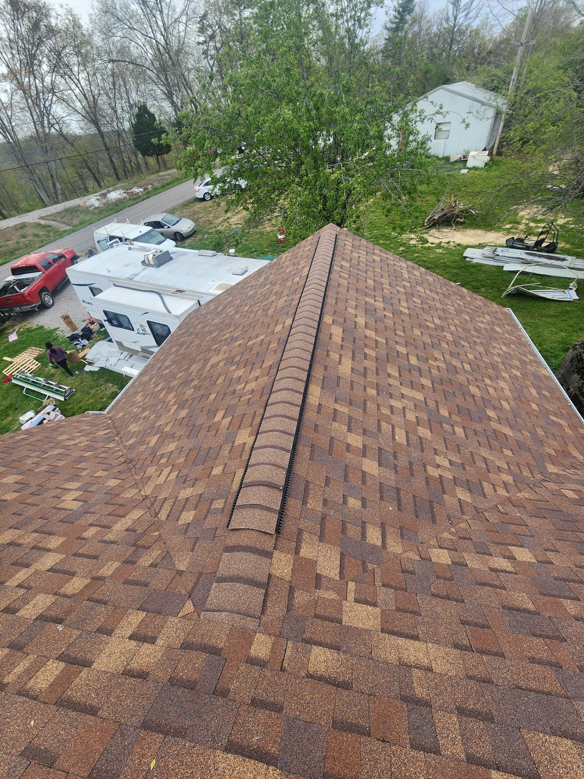 Brown shingle roof with a central ridge, view from above. Surrounding green trees, house, and supplies.