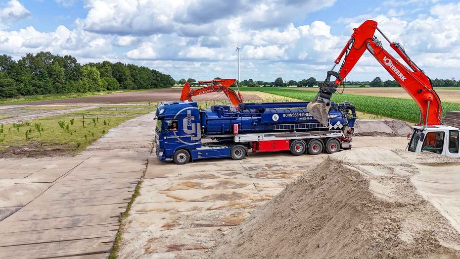 Ein blauer Saugtankwagen steht neben einem roten Bagger, der Sand lädt.