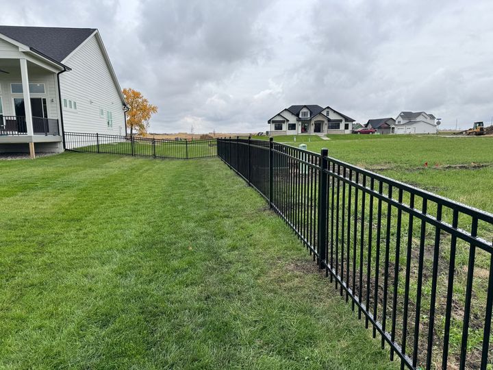Black metal fence along a sidewalk, with a house and trees in the background. 