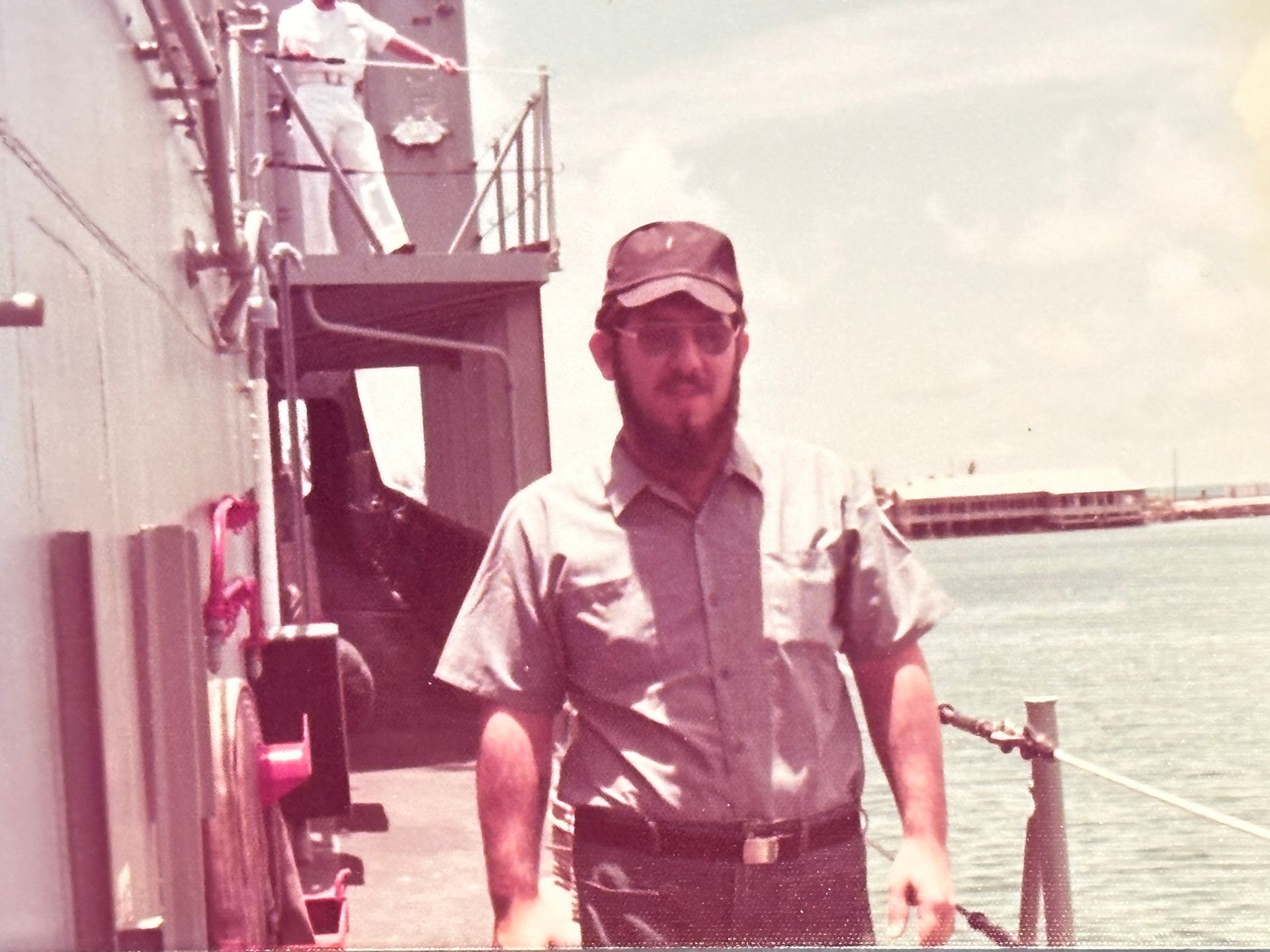 Man in navy uniform on ship deck, looking towards the viewer, wearing a cap. Another figure in white on the deck above.