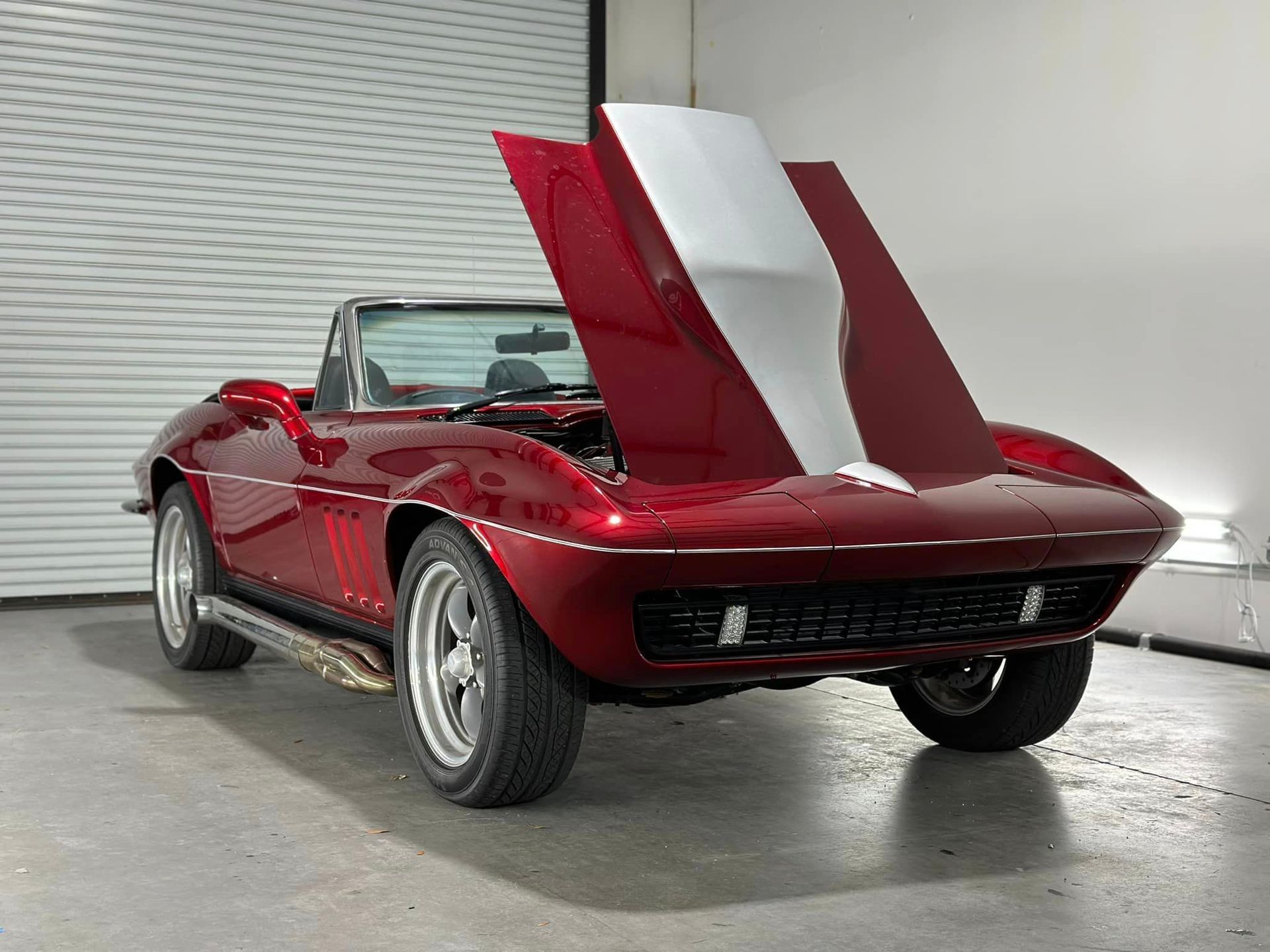 A red corvette convertible with the hood up is parked in a garage.