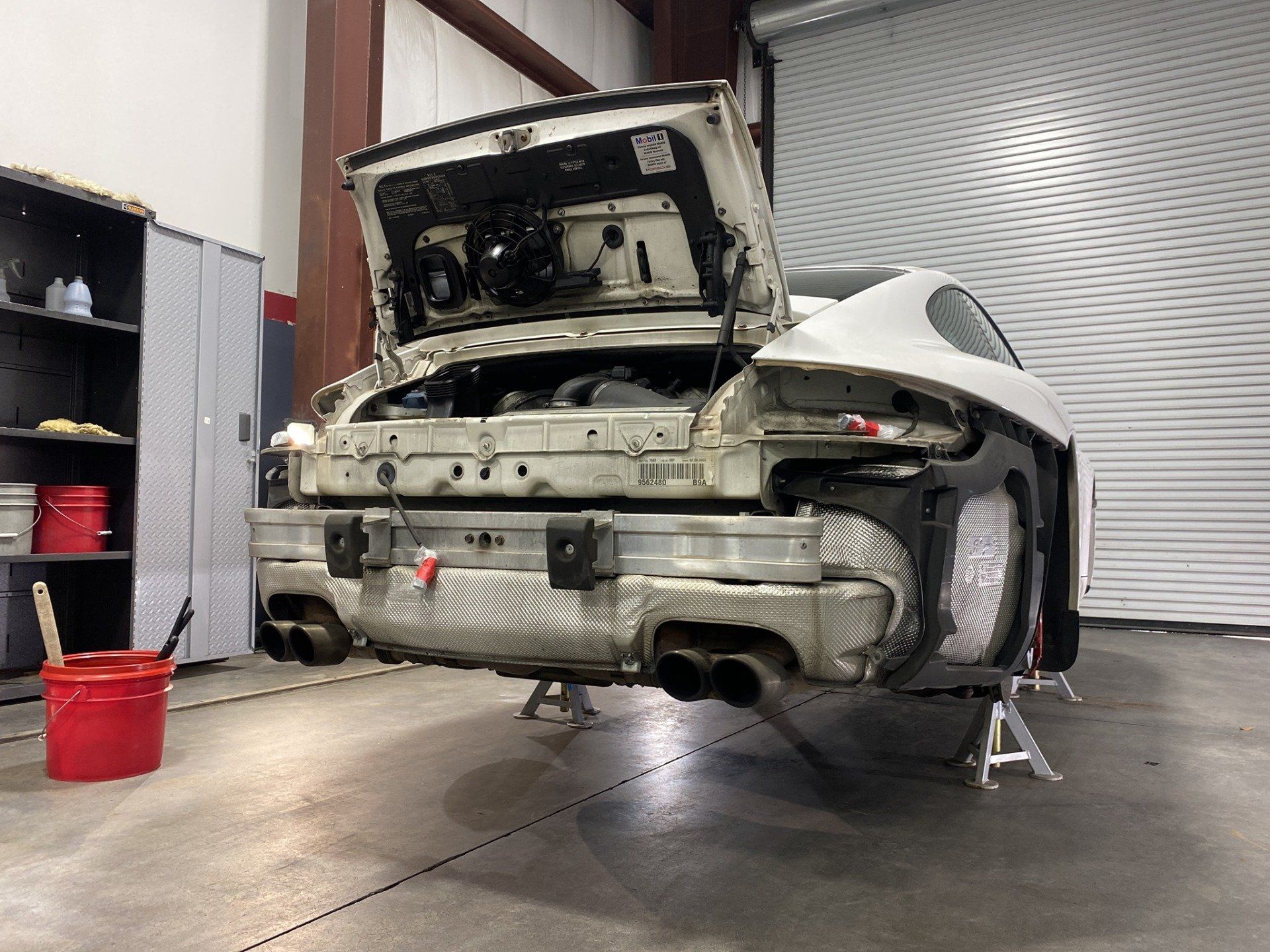 A white car with the hood up is sitting on a stand in a garage.