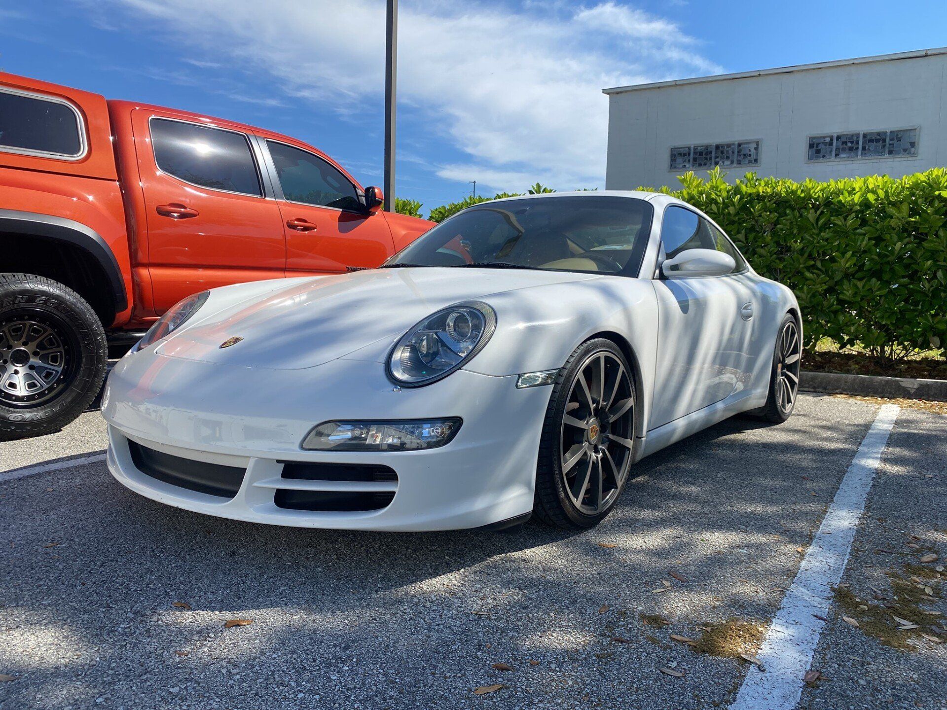 A white porsche 911 is parked in a parking lot next to a red truck.