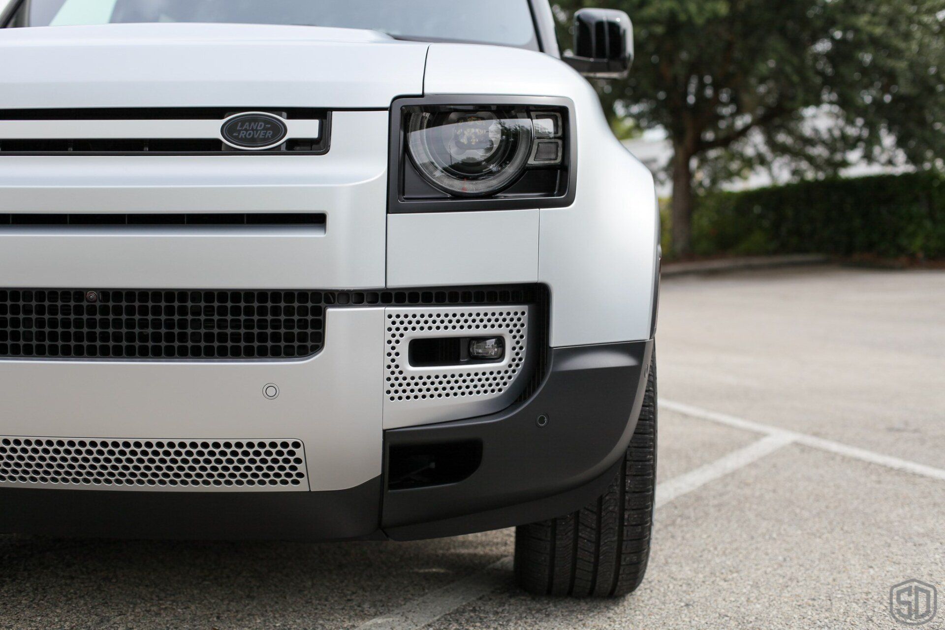 The front of a silver land rover defender is parked in a parking lot.