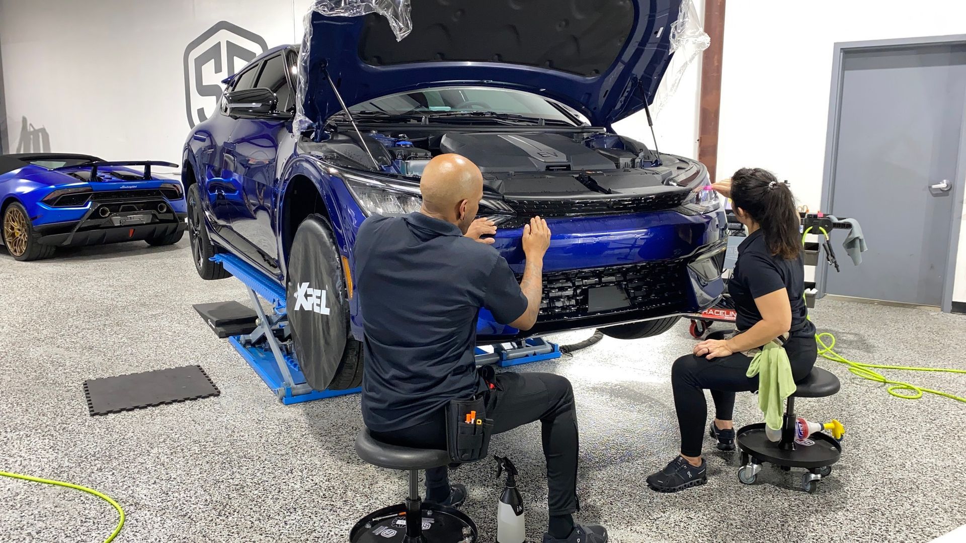 A man and a woman are working on a blue car in a garage.