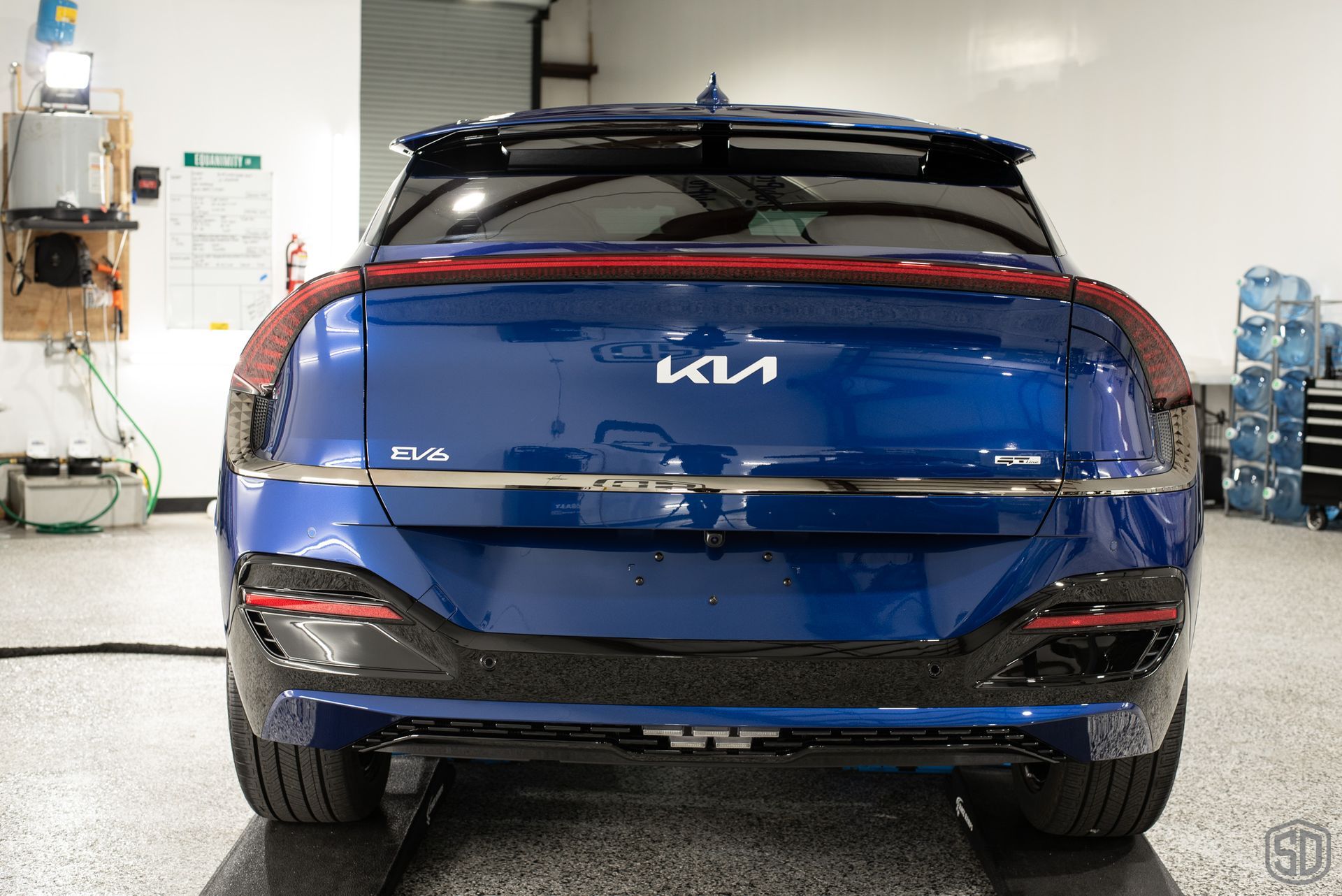 A blue car is parked in a garage with a lot of water bottles in the background.