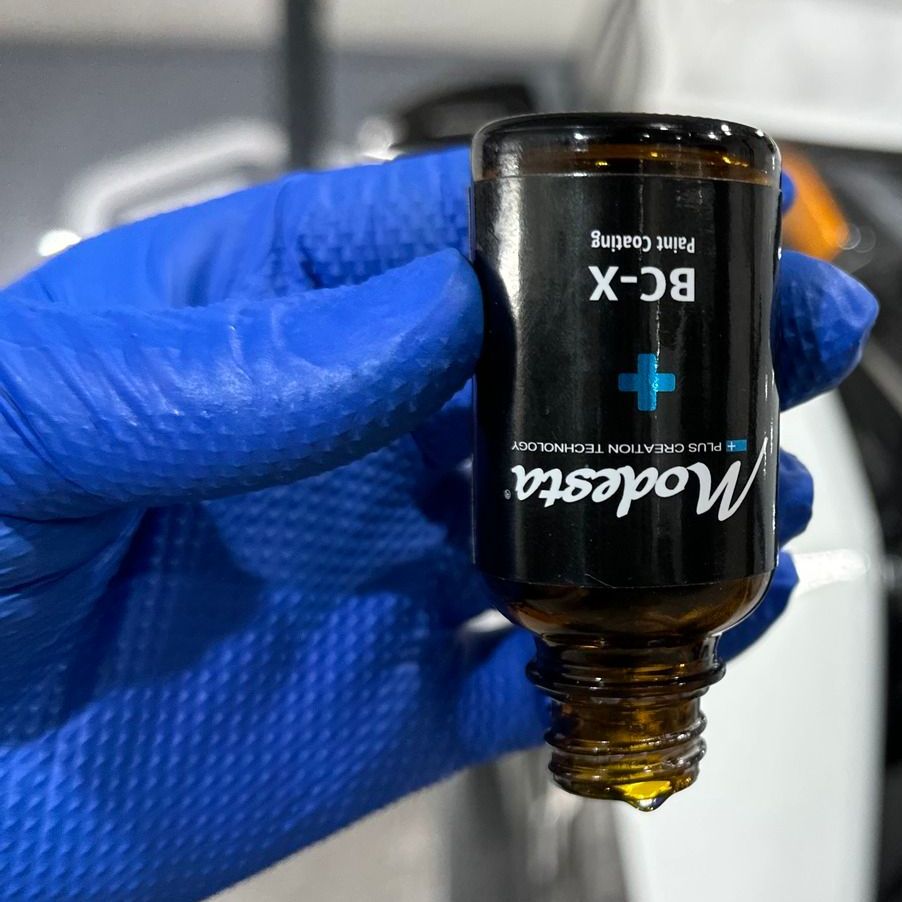 A hand wearing blue gloves is holding a bottle of modesta bc-x paint coating