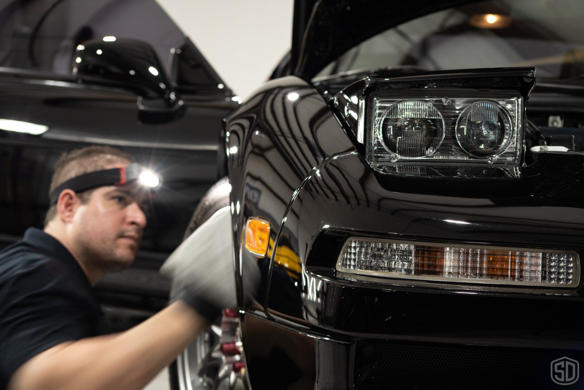 A man wearing a headlamp is working on the front of a black car.