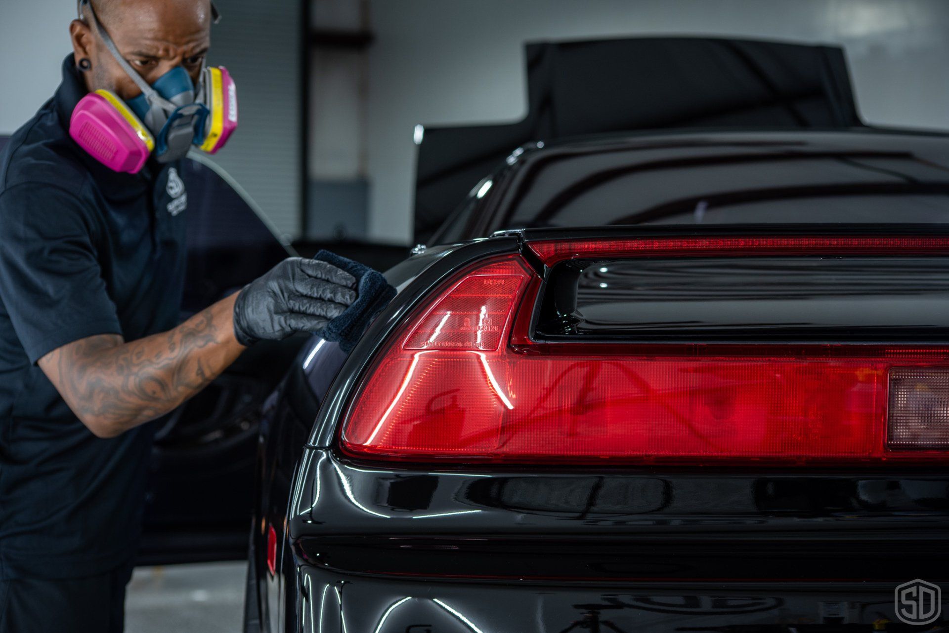 A man wearing a mask is polishing the tail light of a black car.