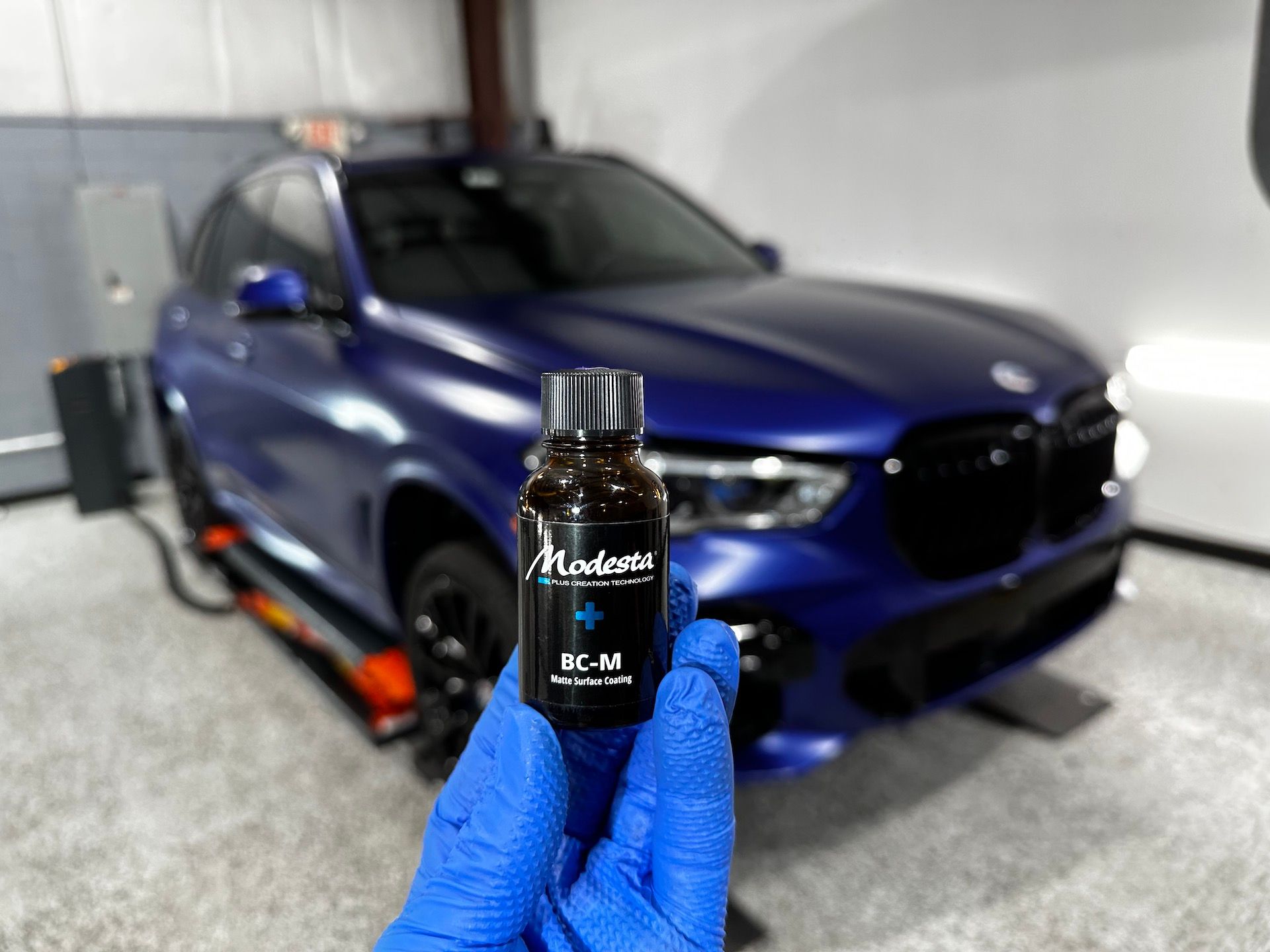 A person is holding a bottle of ceramic coating in front of a blue car.