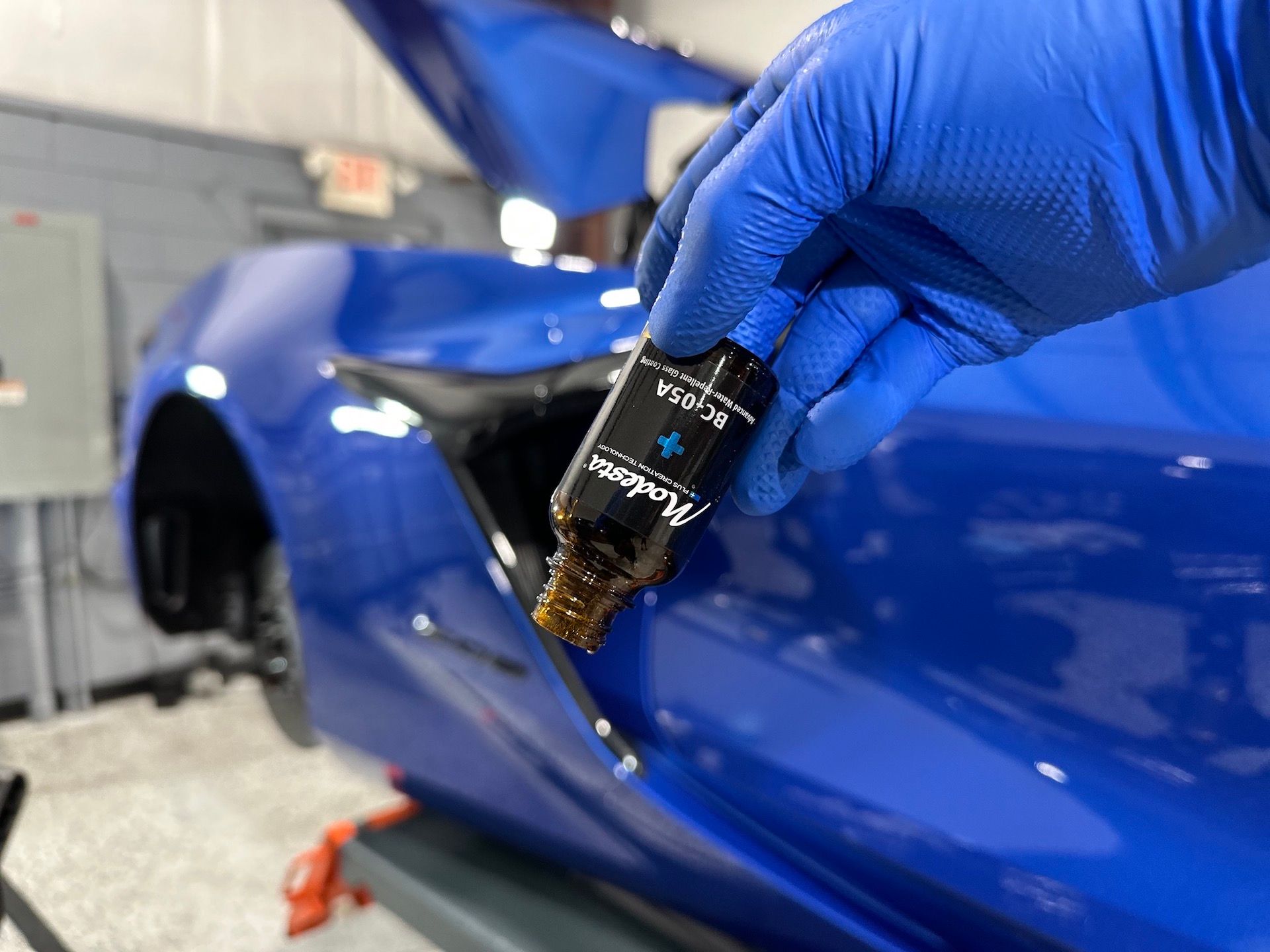 A person wearing blue gloves is holding a bottle of oil in front of a blue car.