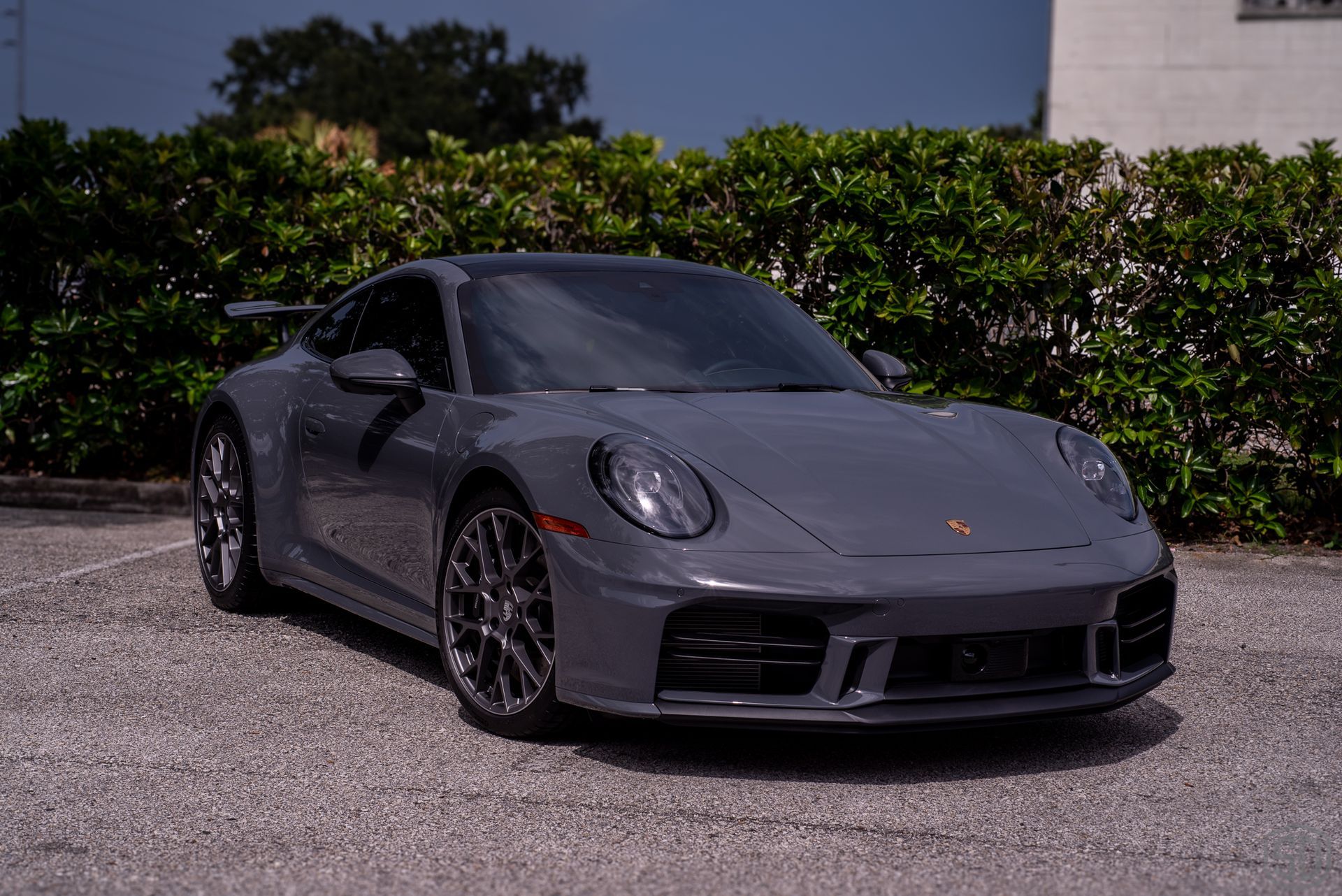 Gray Porsche 911 sports car parked on gravel, in front of green bushes, under a cloudy sky.