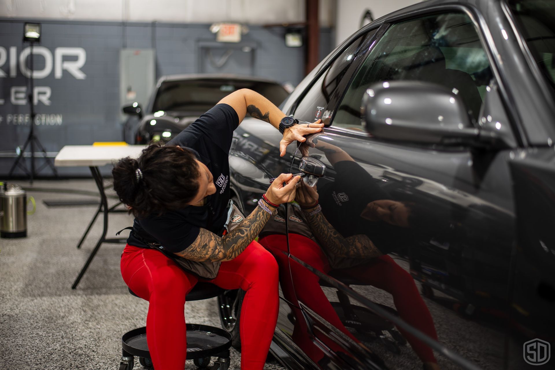 A woman is sitting on a stool working on a car in a garage.