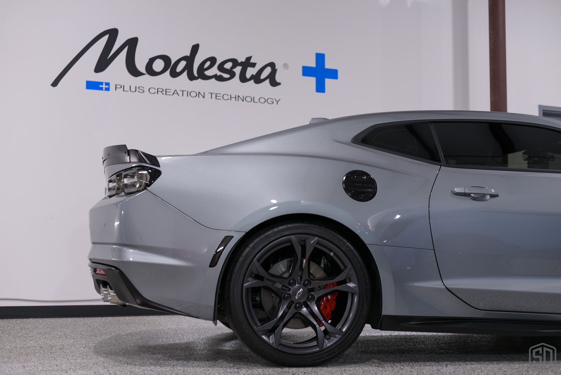 A gray sports car is parked in front of a wall that says modesta on it.