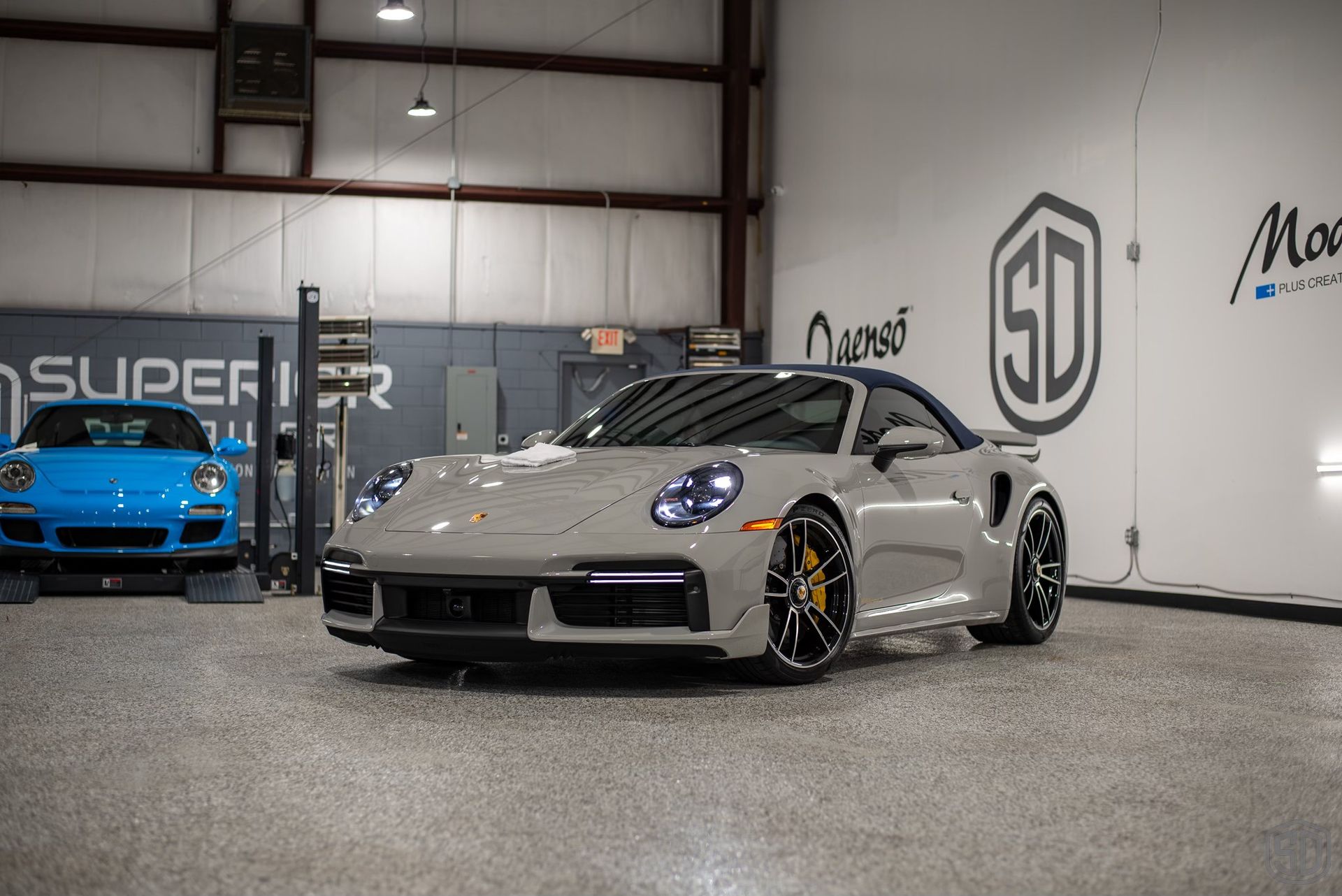 A gray porsche 911 turbo s is parked in a garage next to a blue porsche 911 turbo s.