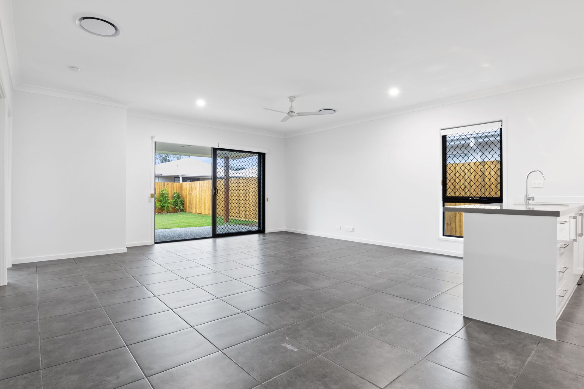 Empty modern living room with gray tile floor, white walls, sliding glass door, and kitchen island on the right