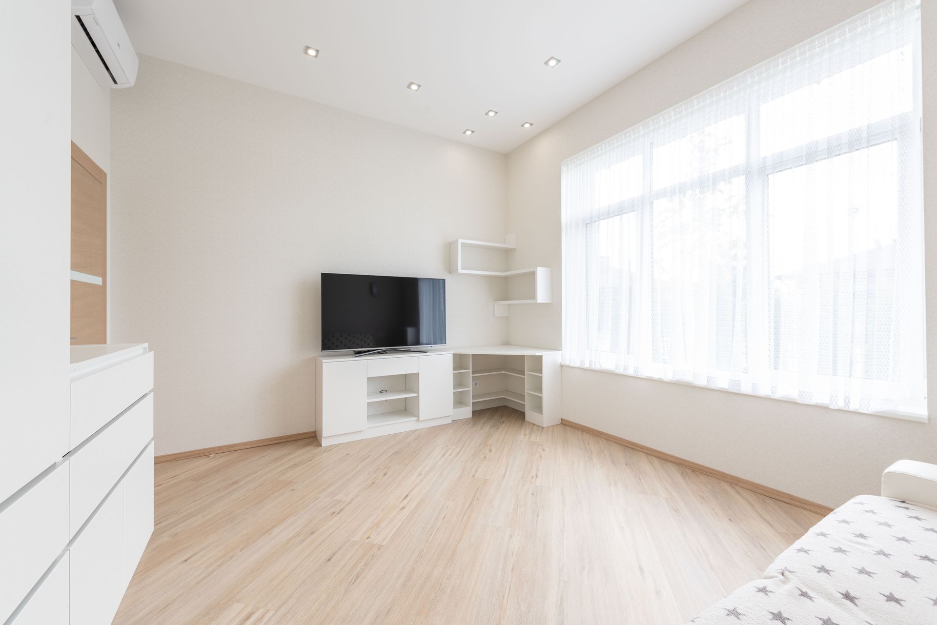 Bright minimalist living room with TV, white furniture, light wood floor, and large sunlit window