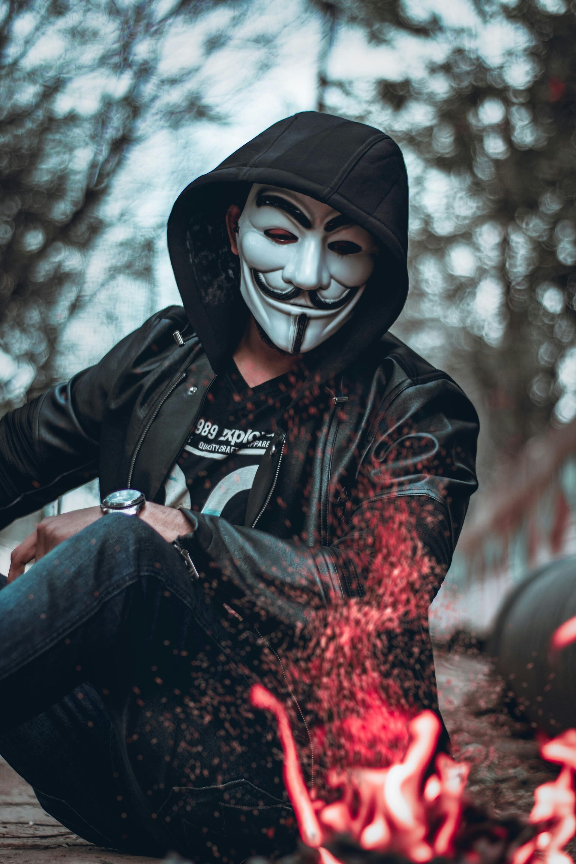 Person wearing a Guy Fawkes mask and hood, near a small fire.