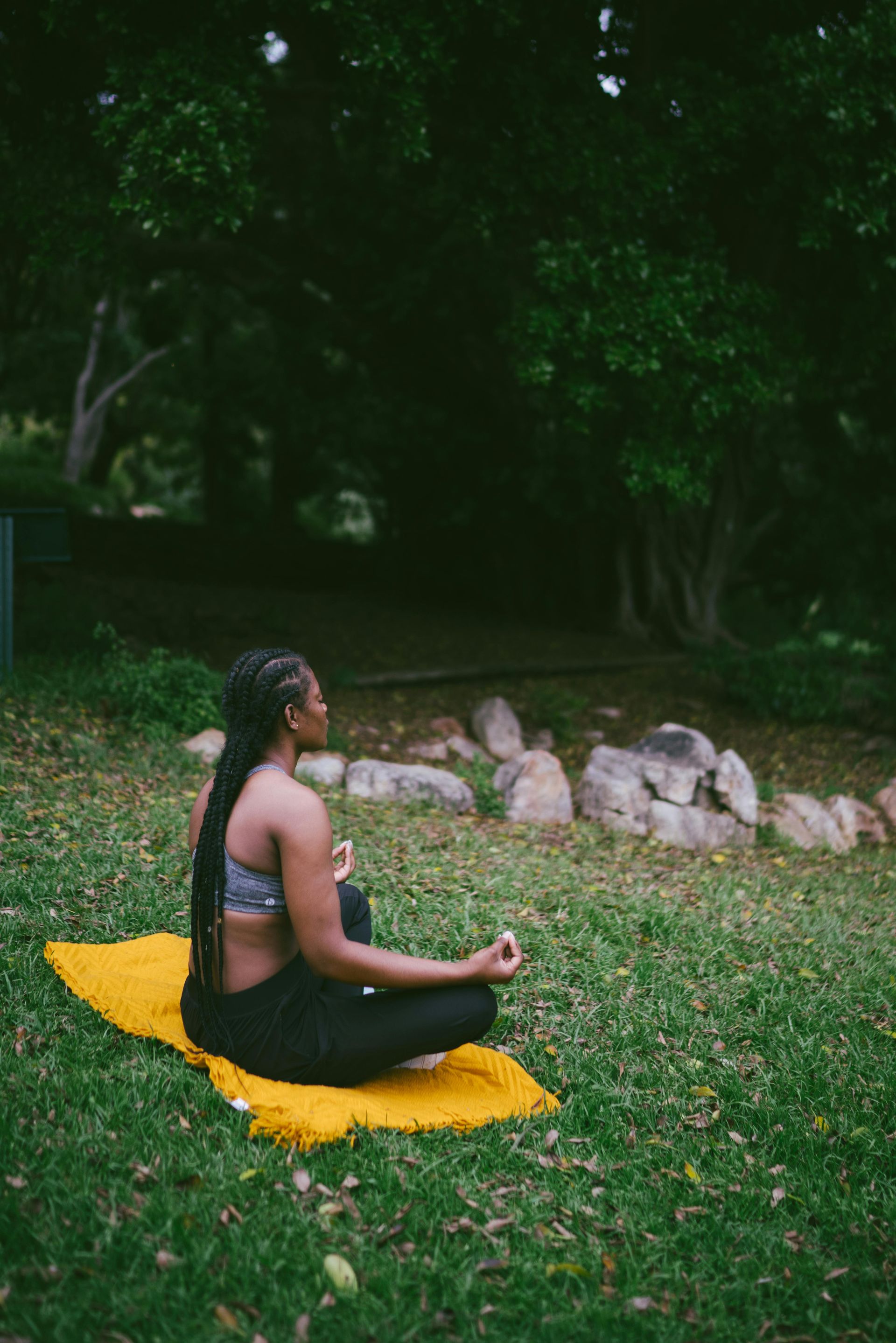 Woman meditating in a park, sitting cross-legged on a yellow mat, hands in prayer pose, surrounded by greenery.
