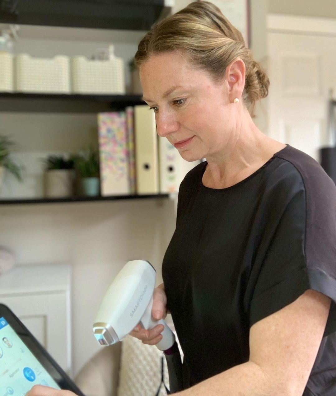 A woman in a black shirt is holding a device and looking at a tablet.