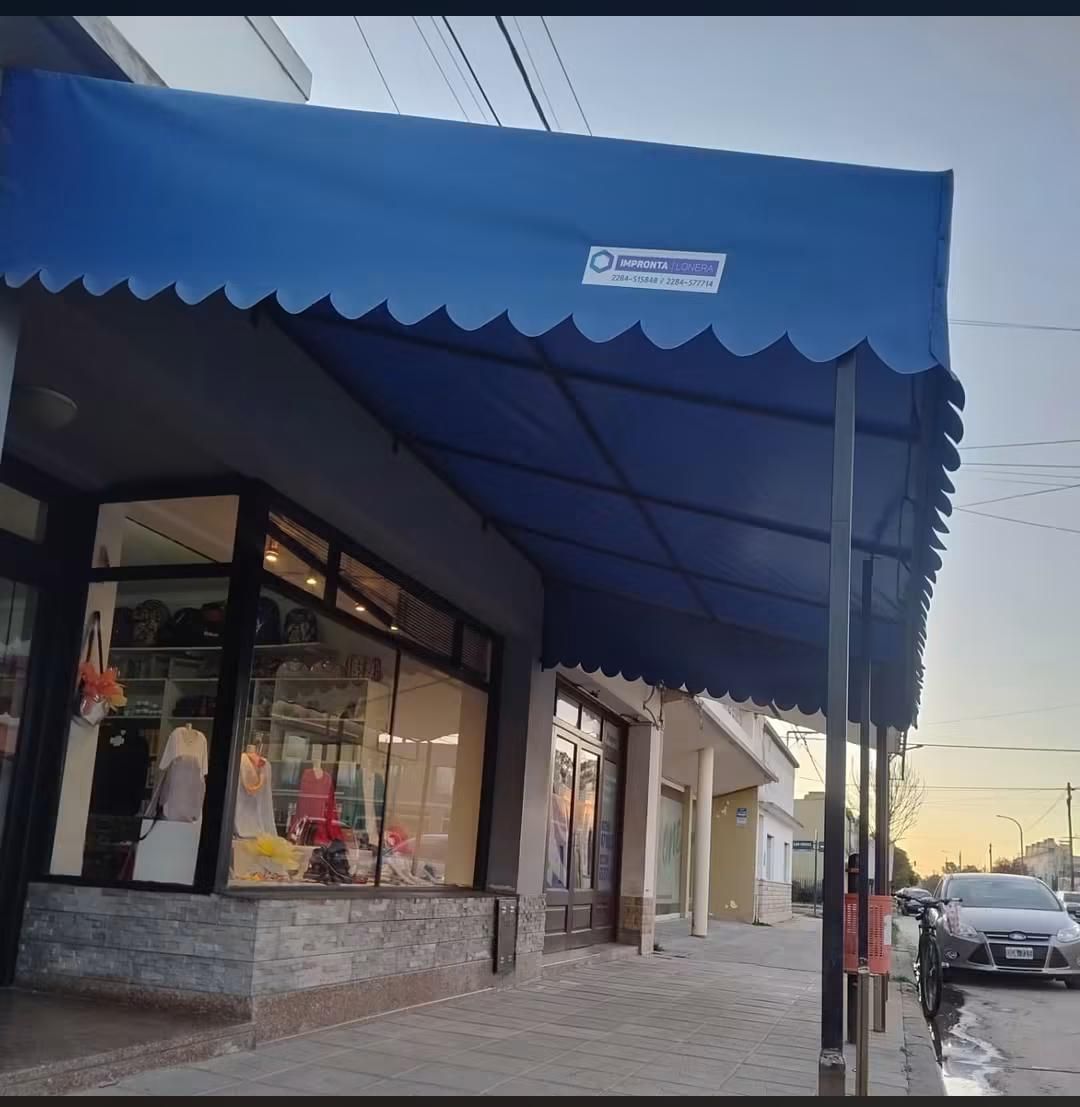 Fachada de una tienda con toldo azul en una acera al anochecer, con grandes ventanales y coches aparcados cerca.