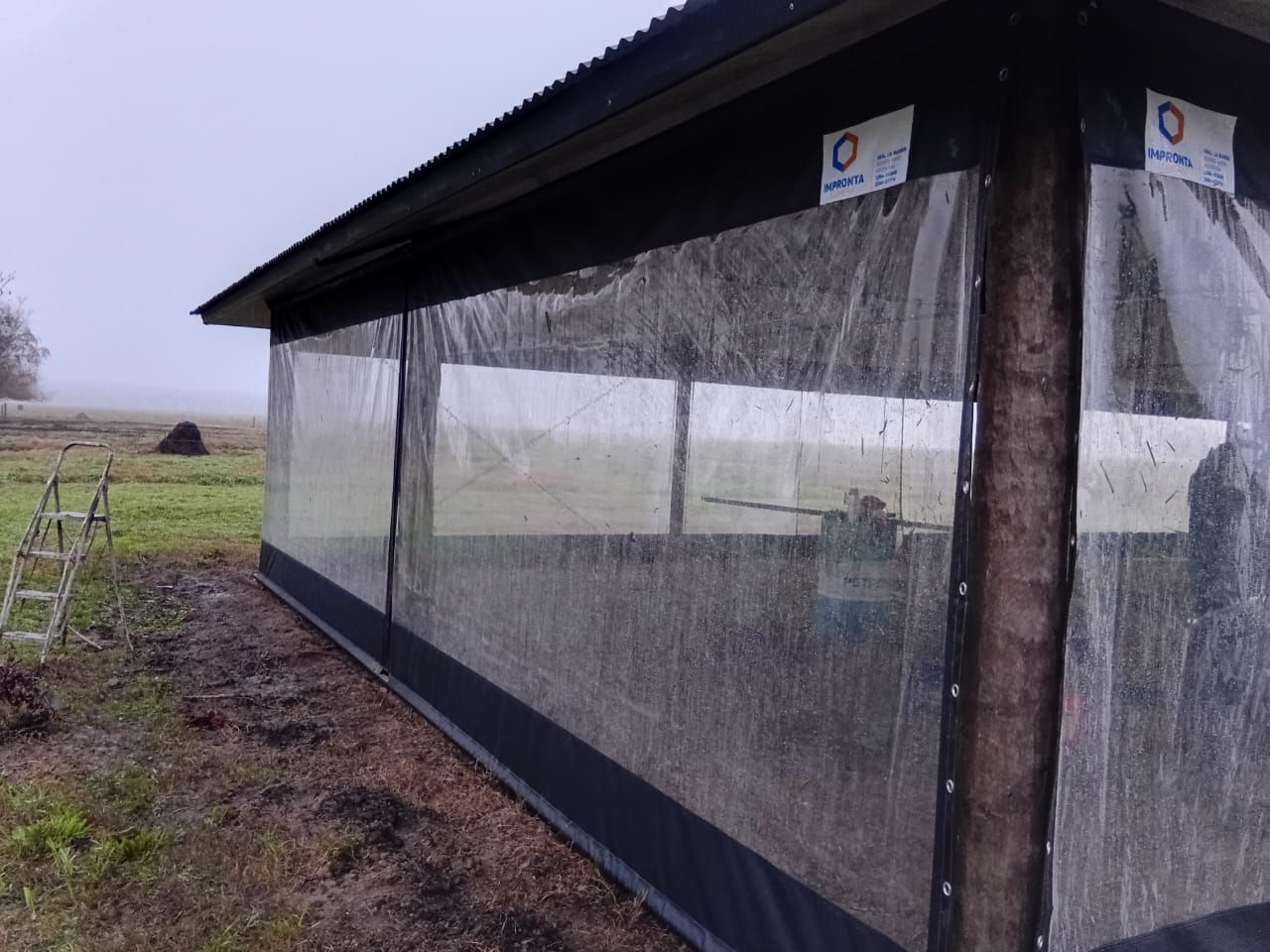 Refugio exterior para la lluvia con techo negro y paneles laterales de plástico transparente junto a un campo de hierba.