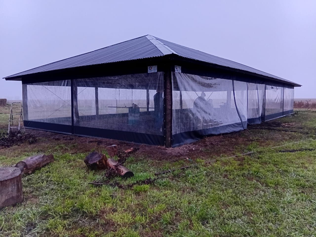 Gran cobertizo negro de malla para ganado en un campo cubierto de hierba y con niebla, con techo de metal