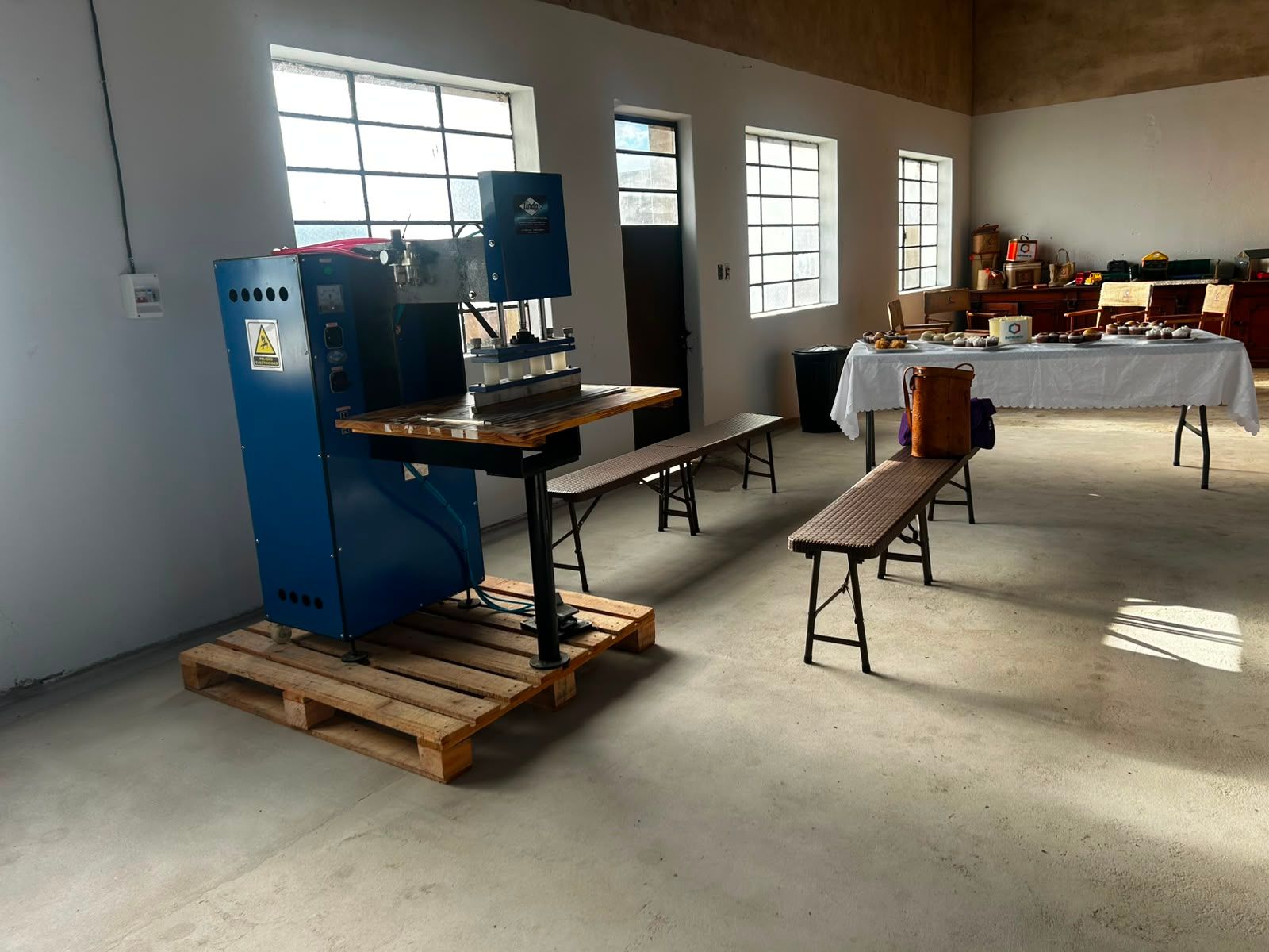 Máquina industrial azul sobre un palé en un taller, con bancos y mesas en una habitación soleada.