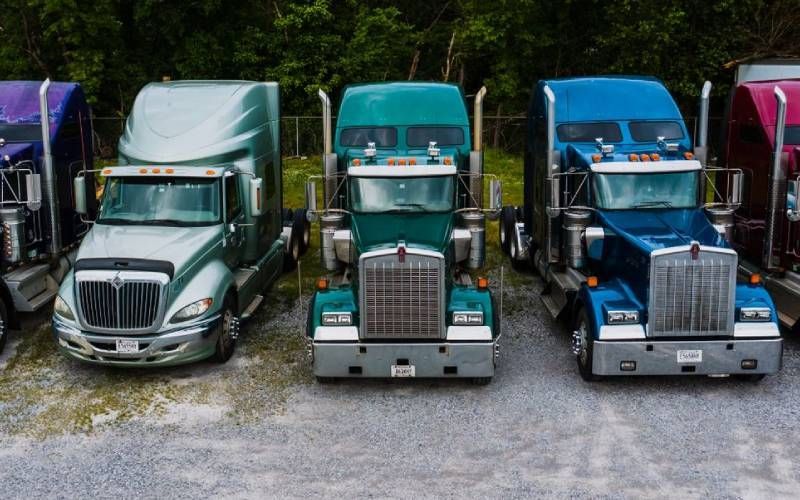 Tres camiones semirremolques estacionados uno al lado del otro sobre grava, con cabinas verdes y azules.