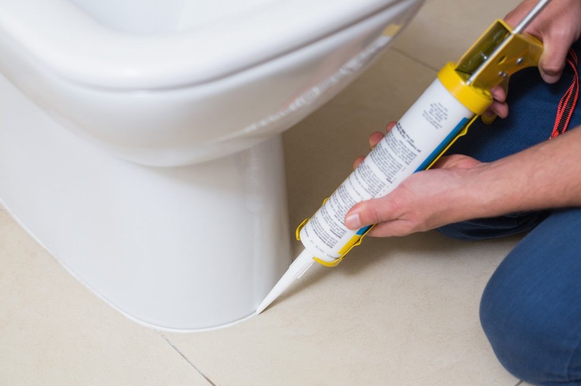 Person applying white caulk to the base of a white toilet on a light-colored tile floor.
