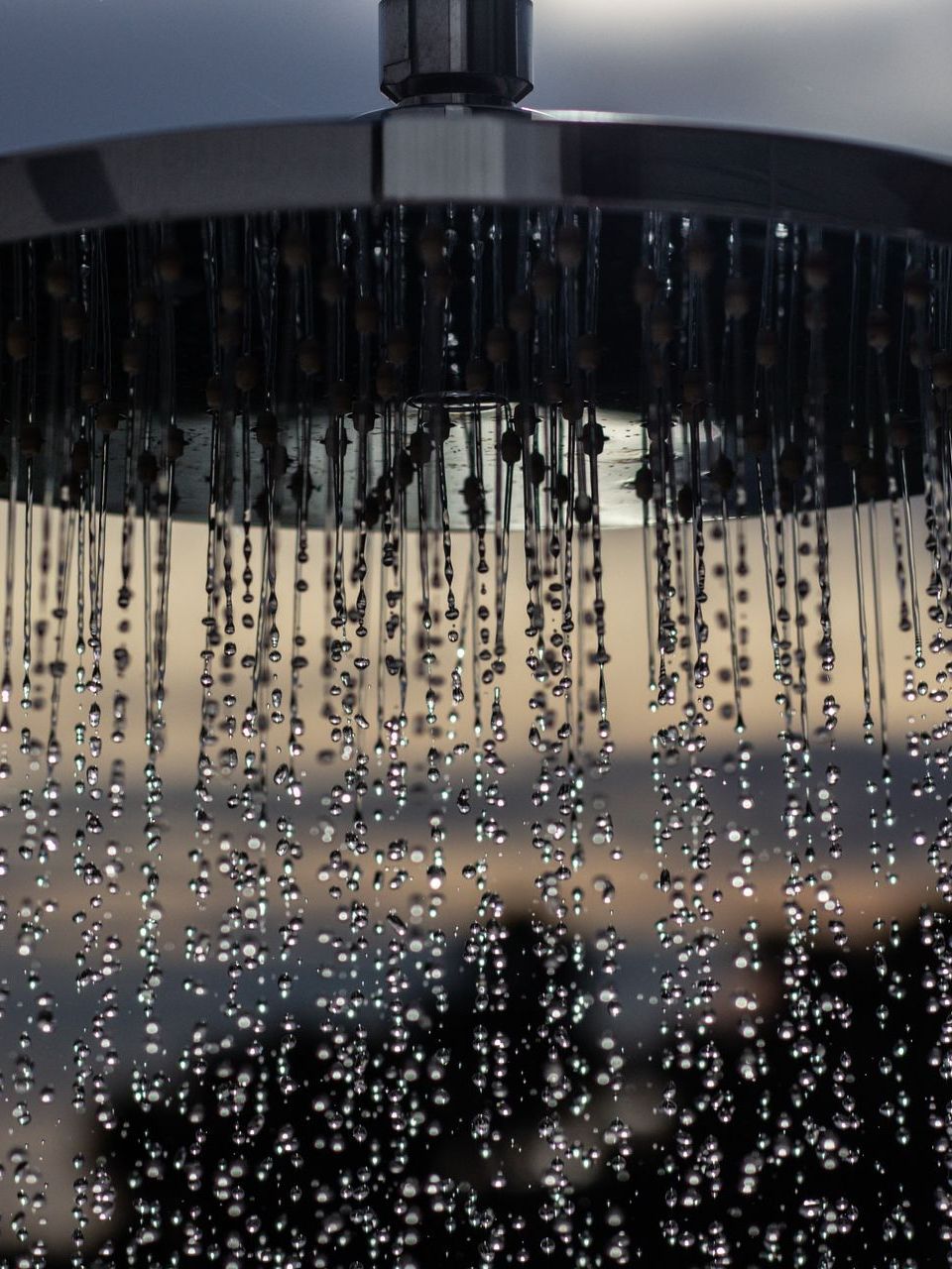 Close-up of a shower head with water droplets falling against a blurry, light-filled background.