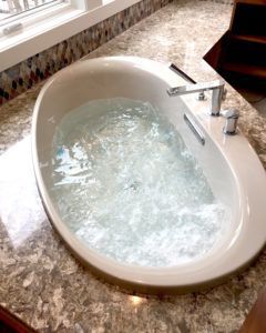 Oval bathtub filled with bubbly water, chrome faucet, granite countertop, near a window.