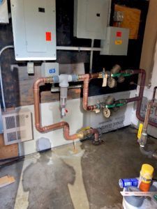 Copper pipes and electrical boxes on a wall, with water on the floor, indicating a leak.