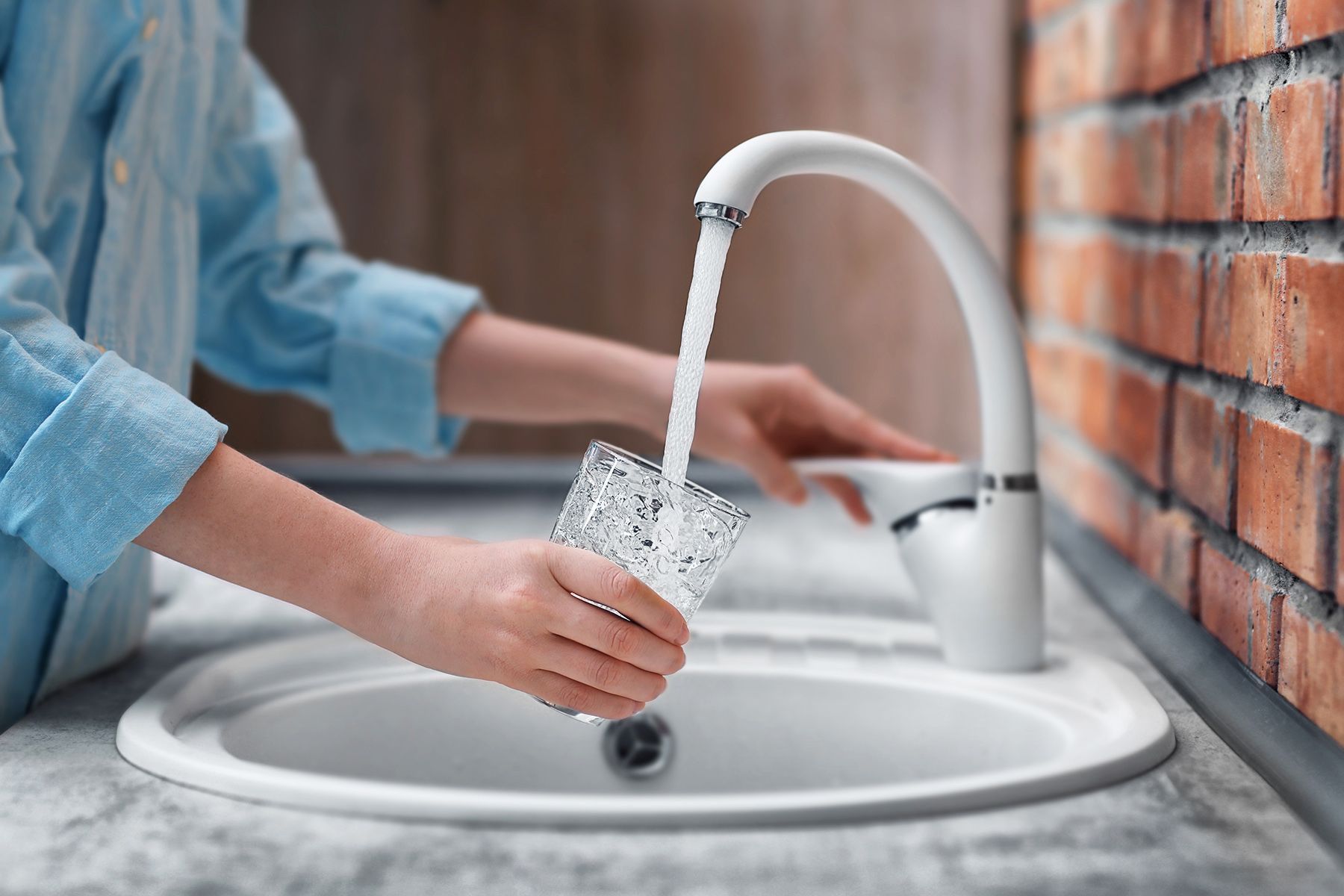 Person filling a glass with water from a white faucet in a sink; brick wall in background.