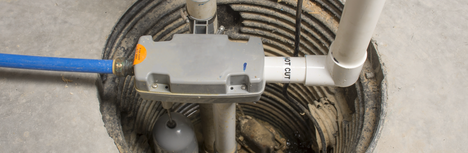 Sump pump system with a gray box, blue and white pipes, installed in a concrete floor.