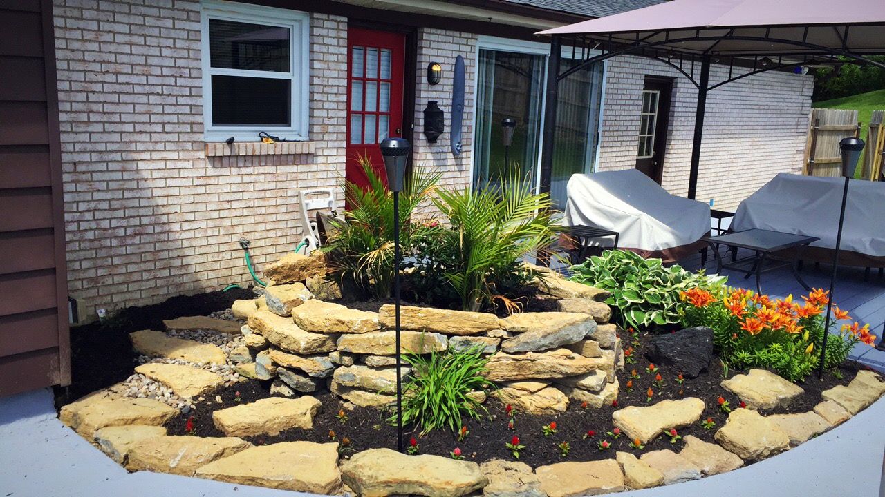 Stone-edged flower bed with plants, mulch, and tiki torches. Patio with covered furniture and a brick building.