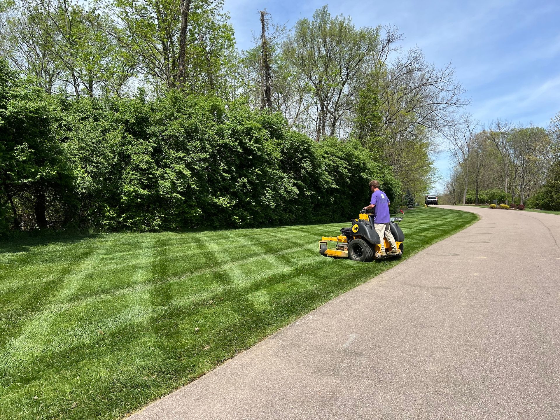 A person mowing a patterned lawn with a yellow riding mower next to a curved road and a green hedge.