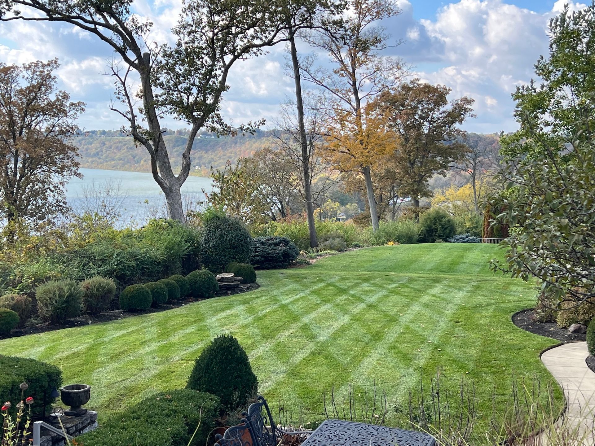 Lush green lawn with striped mowing pattern, overlooking a lake. Trees frame the view, under a partly cloudy sky.
