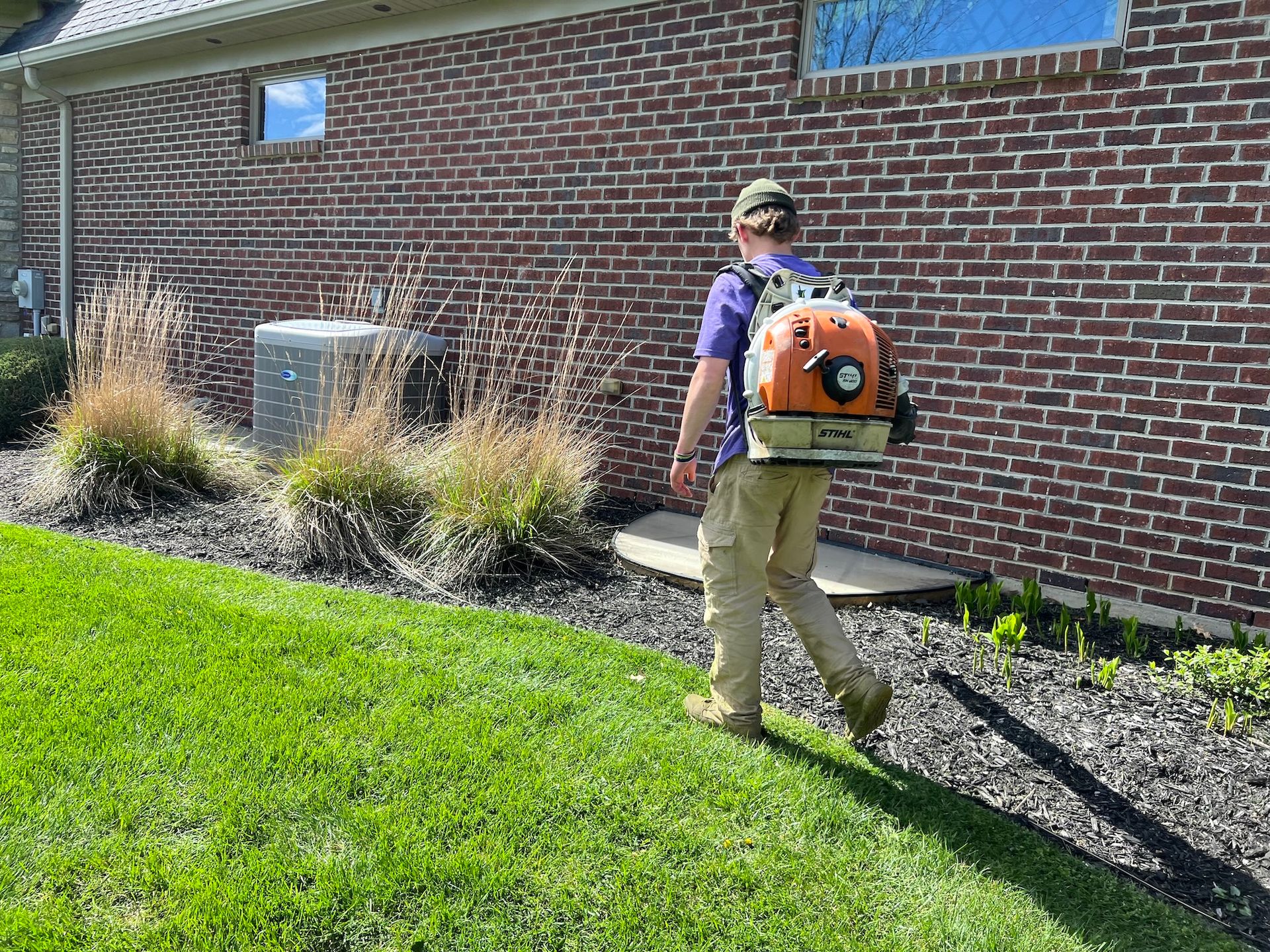 Person with backpack leaf blower walking on grass next to a brick wall.