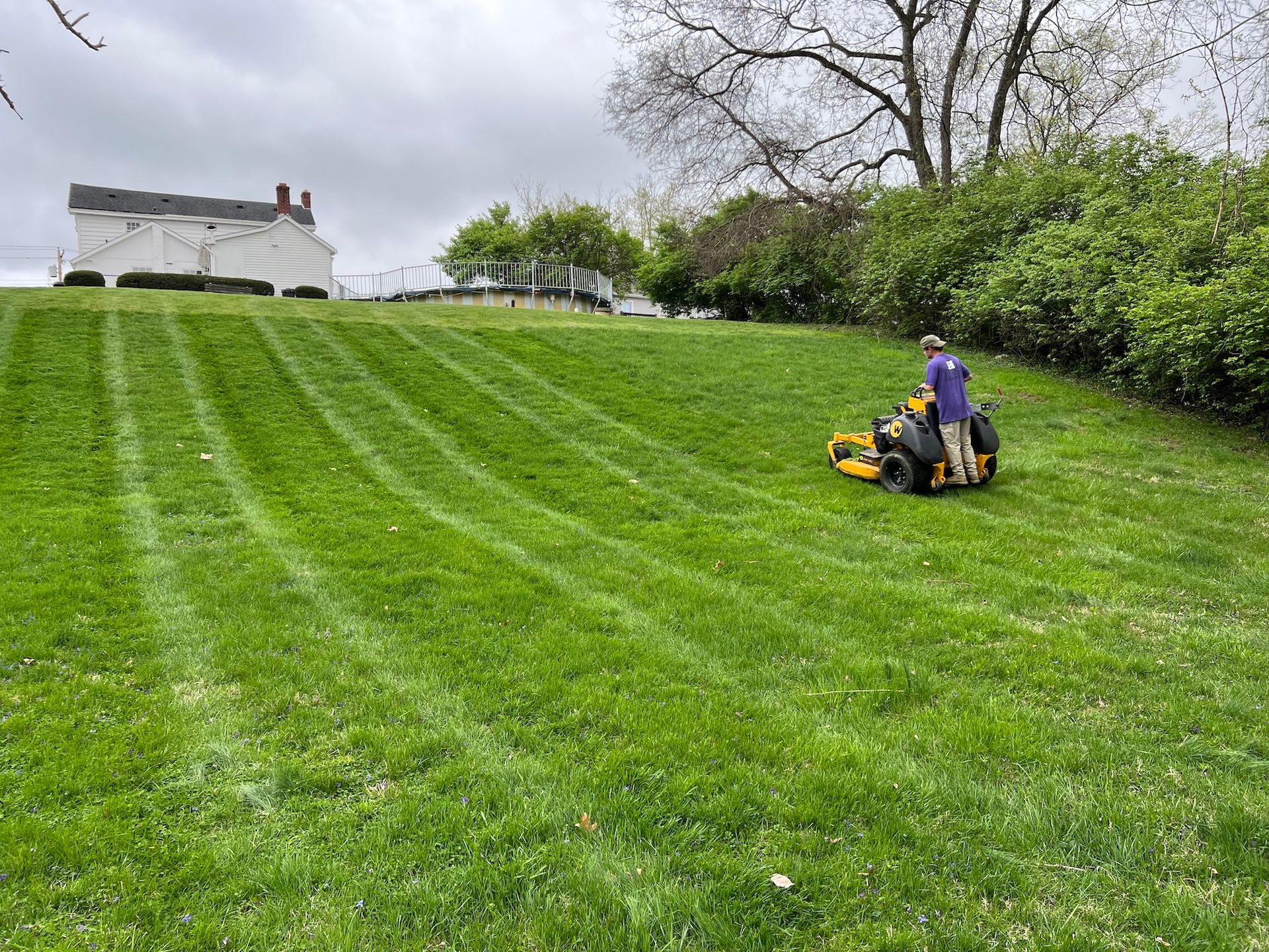 Man mowing a sloped lawn with a riding mower, leaving striped patterns. Green grass, cloudy sky.