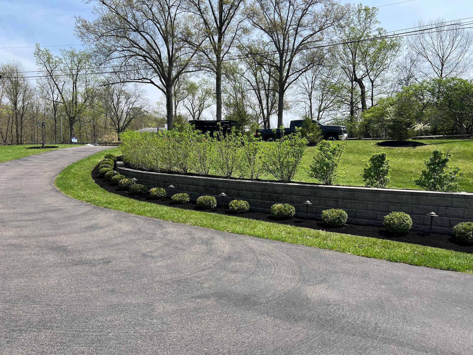 Curving asphalt driveway bordered by a stone retaining wall and manicured bushes, grassy area, trees in the background.