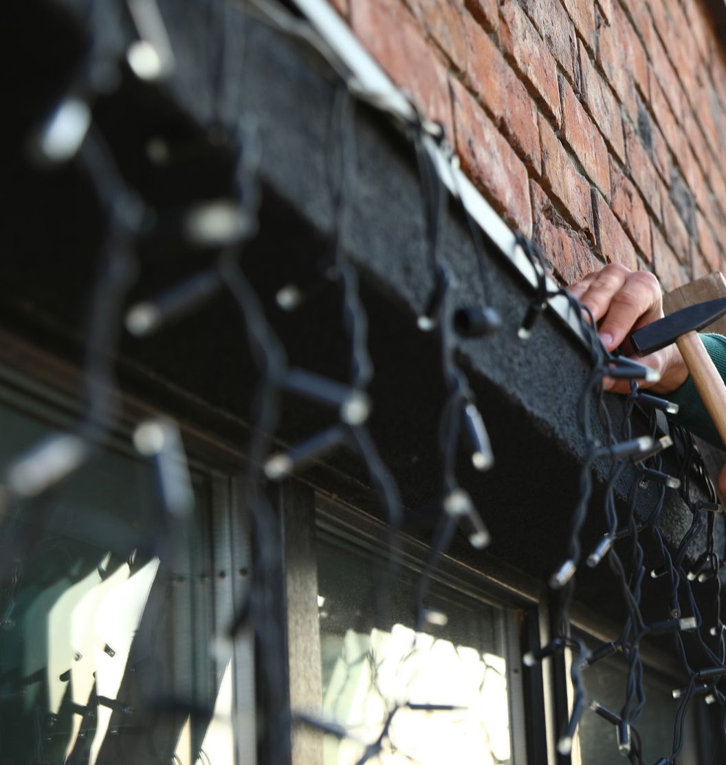Person hanging Christmas lights along a dark eave of a brick building.