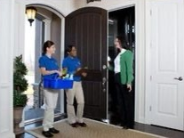 Two people in blue shirts deliver supplies to a woman at a doorway.